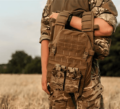Soldier holding tactical plate carrier vest with MOLLE pouches in a field, designed for military protection and gear attachment.
